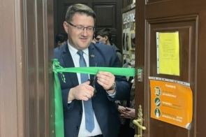Mateusz Pindel Prezes WFOŚiGW w Katowicach przecina zieloną wstęgę przy wejściu do sali. W tle widoczne są osoby stojące w pomieszczeniu, a na drzwiach znajdują się kartki informacyjne.