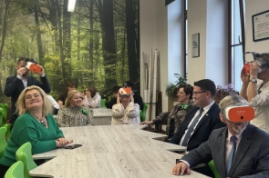 Osoby siedzą przy długim stole w pracowni z zielonymi krzesłami. Część uczestników korzysta z gogli VR, a w tle widoczna jest fototapeta z motywem lasu oraz duże okna.