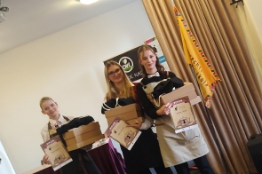 Trzy młode dziewczyny stoją na scenie z dyplomami i nagrodami w rękach, uśmiechając się do zdjęcia. W tle widać sztandar szkoły oraz baner sponsora wydarzenia.