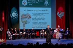 Scena podczas uroczystości. Na pierwszym planie stoi kobieta przy mikrofonie, obok osoba w stroju regionalnym trzyma bukiet kwiatów. W tle widoczne są osoby siedzące na scenie, czerwone kurtyny oraz duży ekran.