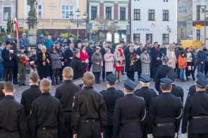 Uczestnicy uroczystości stoją na rynku. Na pierwszym planie widoczne są osoby w mundurach ustawione tyłem, a przed nimi zgromadzeni mieszkańcy z biało-czerwonymi flagami, kwiatami i kotylionami. W tle znajdują się kamienice oraz pomnik.