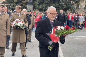 Mężczyzna w czarnym płaszczu trzyma bukiet biało-czerwonych kwiatów. Za nim idą osoby w mundurach z wieńcem, a w tle stoją uczestnicy.