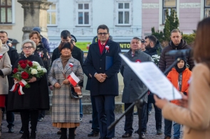 Grupa osób stoi na rynku podczas uroczystości. Część uczestników ma biało-czerwone kotyliony, jedna osoba trzyma bukiet kwiatów, a druga ma małą flagę Polski. Na pierwszym planie widoczny jest mikrofon i kartka trzymana przez osobę przemawiającą.