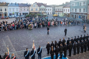 Uroczystość na rynku z udziałem licznie zgromadzonych mieszkańców. Na placu widoczne są poczty sztandarowe, osoby w mundurach oraz uczestnicy stojący przed zabytkowymi kamienicami.