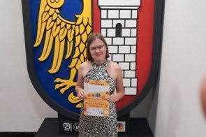 Młoda kobieta w srebrnej sukience stoi przed dużym kolorowym herbem Gliwic, trzymając w dłoniach dyplom oraz pamiątkową teczkę, uśmiechając się do zdjęcia.