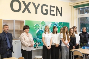 Pracownia z dużym napisem „OXYGEN” na ścianie. W pomieszczeniu stoją uczniowie oraz osoby dorosłe, a w tle widoczne są szafki, mikroskop i grafika z motywem cząsteczek.
