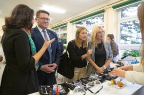 Osoby stoją przy stole z wyposażeniem pracowni. Na blacie widoczne są m.in. okulary ochronne, narzędzia, próbki oraz mikroskop, a w tle znajdują się duże okna i rośliny.