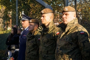 Osoby w mundurach stoją w szeregu podczas uroczystości. Na pierwszym planie widoczni są żołnierze w umundurowaniu polowym, a obok stoi osoba w granatowym mundurze.