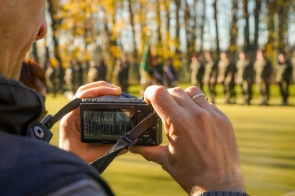 Osoba trzyma aparat fotograficzny i robi zdjęcie uroczystości na boisku. W tle, poza ostrością, widoczna jest grupa umundurowanych osób stojących w szyku.