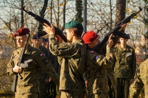 Umundurowane osoby stoją w szyku i trzymają broń. Na pierwszym planie dwie osoby unoszą karabiny, a w tle widoczni są kolejni uczestnicy w mundurach.