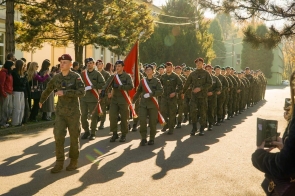 Umundurowane osoby maszerują drogą w zwartym szyku. Na przodzie idzie poczet ze sztandarem, a po bokach stoją obserwujący.