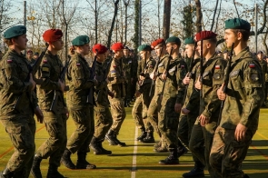 Umundurowane osoby maszerują w szyku na boisku. Część z nich trzyma broń, a na mundurach widoczne są polskie flagi oraz berety w różnych kolorach.