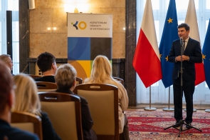 Marek Wócik Wojewoda Śląski stoi przy mikrofonie przed publicznością. W tle widoczne są flagi Polski i Unii Europejskiej oraz baner Kuratorium Oświaty w Katowicach.