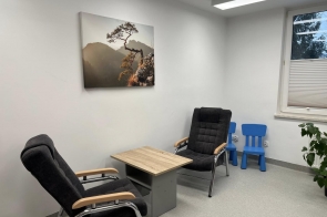 Jasne pomieszczenie po remoncie, wyposażone w dwa fotele, stolik oraz małe krzesełka dla dzieci. Na ścianie wisi obraz z krajobrazem, a przy oknie stoi roślina.