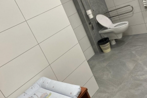 Łazienka w jasnoszarej kolorystyce, wyposażona w toaletę z uchwytami oraz przewijak dla dziecka. W pomieszczeniu widać płytki na ścianach i podłodze, okno oraz grzejnik.