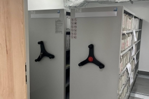 Pomieszczenie archiwum z przesuwnymi regałami na dokumenty. Widać półki z segregatorami i teczkami, szarą podłogę oraz instalacje wentylacyjne pod sufitem.