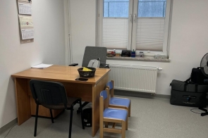 Małe pomieszczenie biurowe w jasnej kolorystyce. Przy oknie stoi biurko z krzesłami, obok znajduje się małe krzesełko dziecięce, a na ścianie wisi kalendarz.