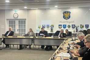 Radni siedzą przy stołach w sali sesyjnej. Na blatach znajdują się mikrofony, tablety, dokumenty, kubki i tabliczki imienne. 
