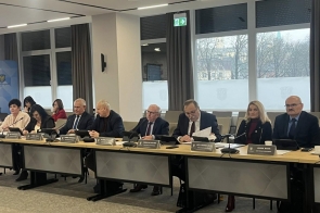 Radni siedzą przy stołach w sali sesyjnej. Na blatach znajdują się mikrofony, tablety, dokumenty, kubki i tabliczki imienne. W tle widoczne są duże okna oraz zasłony.