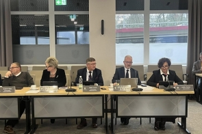 Radni siedzą przy stołach w sali sesyjnej. Na blatach znajdują się mikrofony, tablety, dokumenty, kubki i tabliczki imienne. W tle widoczne są duże okna oraz zasłony.