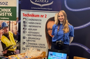  Osoba w niebieskiej koszulce stoi przy banerze ZSEG w Cieszynie, koszu z materiałami promocyjnymi oraz laptopie z prezentacją.