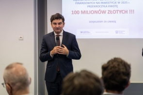Mężczyzna w garniturze stoi z mikrofonem przed publicznością. W tle widoczny jest ekran z prezentacją, na którym wyróżnia się napis „100 milionów złotych”.