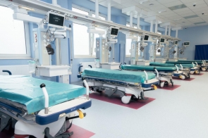 Sala szpitalna w jasnoniebieskiej kolorystyce, wyposażona w kilka łóżek transportowych oraz stanowiska z monitorami i aparaturą medyczną.