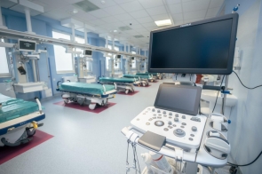 Sala szpitalna w jasnoniebieskiej kolorystyce, wyposażona w łóżka transportowe, aparaturę medyczną oraz urządzenie z monitorem na pierwszym planie.