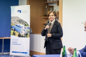 Kobieta stoi z mikrofonem w sali konferencyjnej. Obok znajduje się roll-up Szpitala Śląskiego w Cieszynie, a na stole widoczne są butelki z wodą.