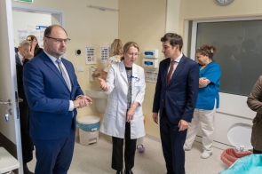 Pomieszczenie szpitalne w jasnej kolorystyce. Lekarka w białym fartuchu rozmawia z dwoma mężczyznami w garniturach, a w tle znajdują się osoby z personelu medycznego.