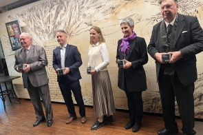 Pięć osób pozuje do zdjęcia, trzymając wyróżnienia z napisem „Cieszynit 2025”. W tle widoczna jest ściana z grafikami i tekstami, a uczestnicy stoją w eleganckich strojach.