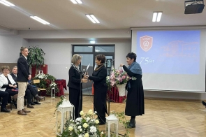 Kilka osób stoi w sali szkolnej przy mikrofonach. Jedna z kobiet trzyma bukiet kwiatów, a w tle widać ekran z napisem „75 lat” oraz dekoracje kwiatowe.