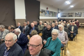 Publiczność siedzi na krzesłach w sali szkolnej. W tle widać ściany z tablicami pamiątkowymi i zdjęciami klasowymi.