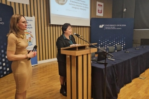 Na zdjęciu widać dwie kobiety. Jedna przemawia przy mównicy, druga stoi obok z mikrofonem. W tle widoczne są banery uczelni i stowarzyszenia oraz ekran z prezentacją.