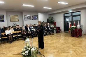 Kobieta stoi przy mikrofonie w sali szkolnej i trzyma teczkę. W tle widać siedzącą publiczność, dekoracje kwiatowe oraz flagi.