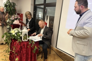 Kilka osób znajduje się przy stole z dokumentami i dekoracjami kwiatowymi. Jedna osoba siedzi i podpisuje dokument, a obok stoją pozostali uczestnicy (podpisanie porozumienia z Hutą Trzyniec).