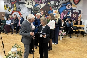 Kilka osób stoi w sali szkolnej i trzyma teczki z dokumentami. W tle widać siedzącą publiczność oraz ścianę z kolorowym muralem.