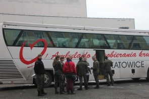 Przy białym autobusie mobilnego punktu pobierania krwi stoi grupa osób w wojskowych mundurach oraz jedna osoba w czerwonej kurtce. Autobus ma duże okna i czerwone oznaczenia związane z krwiodawstwem. Część osób rozmawia i trzyma dokumenty, a do pojazdu prowadzą otwarte drzwi.