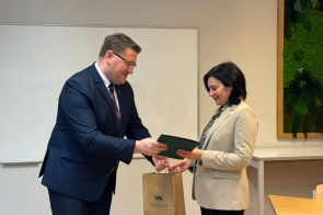 Prezes WFOŚ w ciemnym garniturze podaje Dyrektorce szkoły w jasnej marynarce zieloną teczkę, a w drugiej ręce trzyma papierową torbę z logo. Kobieta odbiera teczkę, uśmiechając się lekko. Scena rozgrywa się w jasnym pomieszczeniu z tablicą na ścianie oraz zieloną dekoracją roślinną w ramie.