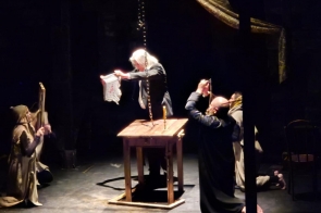 Aktorzy występują na scenie w ciemnej, teatralnej scenografii. W centrum znajduje się drewniany stół ze świecą, a postacie w kostiumach odgrywają scenę przy skupionym świetle.
