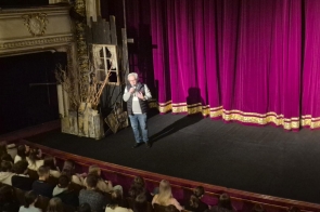 Mężczyzna stoi na scenie teatralnej i przemawia do publiczności. W tle widać purpurową kurtynę oraz elementy scenografii, a na widowni siedzi młodzież.