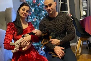 Rodzina pozuje do zdjęcia przy udekorowanej choince. Kobieta trzyma niemowlę, mężczyzna siedzi obok z psem. Zdjęcie wykonano w domowym, świątecznym otoczeniu.