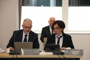 Dwie osoby siedzą przy stole z mikrofonami, laptopem, tabletem i tabliczkami imiennymi. W tle przy oknie siedzi mężczyzna.