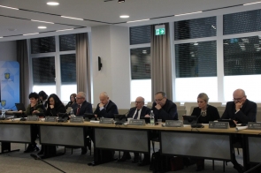 Osoby siedzą przy stołach ustawionych w sali sesyjnej.Przed nimi znajdują się mikrofony, dokumenty, tablety i tabliczki imienne, a w tle widać duże okna oraz opuszczone rolety.