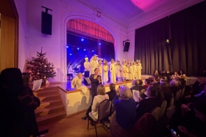 Dzieci w jasnych strojach występują na scenie przed publicznością. Obok sceny stoi choinka, a sala jest oświetlona kolorowym światłem scenicznym.