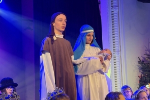Dzieci w strojach jasełkowych stoją na scenie. Jedno z nich trzyma lalkę niemowlęcia, a wokół siedzą inne dzieci w przebraniach i koronach. W tle widać niebieskie oświetlenie sceniczne oraz choinkę.