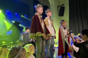 Dzieci w koronach i pelerynach stoją na scenie wśród innych dzieci w przebraniach. Jedno z dzieci trzyma mikrofon, a w tle widać kolorowe oświetlenie sceniczne oraz fortepian.