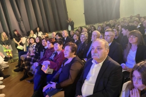 Publiczność siedzi na widowni w sali z niebieskim oświetleniem. W pierwszych rzędach widać dorosłych i młodzież.