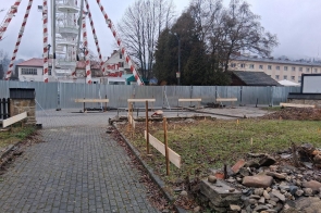 Alejka z kostki brukowej prowadząca przez teren prac budowlanych. Po bokach trawniki z kamieniami i drewnianymi zabezpieczeniami. W tle metalowe ogrodzenie, zabudowania oraz widoczna konstrukcja diabelskiego młyna.