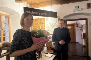 Kobieta trzyma bukiet kwiatów w sali muzealnej. Obok niej stoi mężczyzna, a w tle widać  drewniane elementy wnętrza.